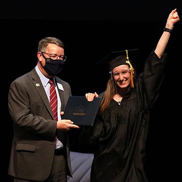 一个带着帽子的高中毕业生, 身穿长袍、戴着面罩的她接受了毕业证书，对着镜头微笑，并在空中举起了拳头