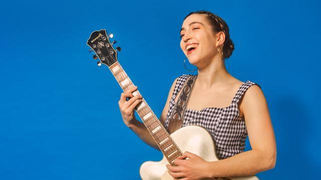Alisa Amador一边唱歌一边弹吉他. 她穿着格子布连衣裙，站在明亮的蓝色背景前.