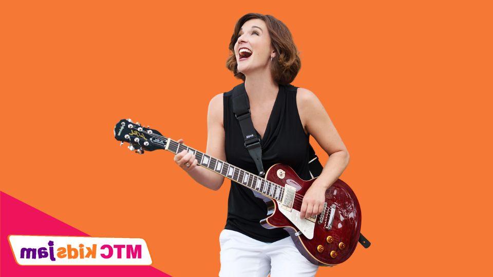 Krista Tatschl Eyler smiles while holding a guitar. She is standing against an orange background.