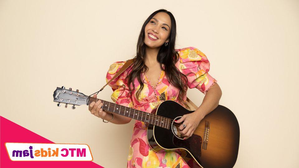 Sonia De Los Santos stands against a beige background, and smiles while holding a guitar. She is wearing a floral dress.