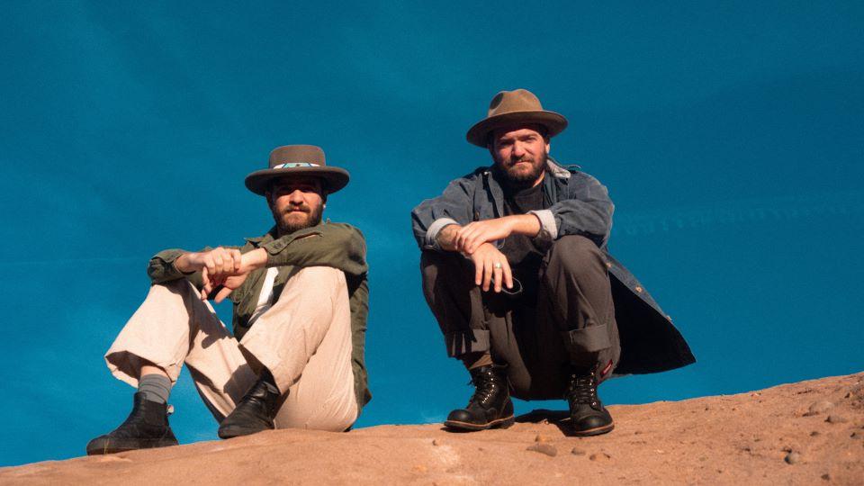 Upward looking picture of two men on top of a dirt hill with the blue sky in the background. One is squatting and one is sitting; both are looking down toward the camera and both are wearing cowboy hats.